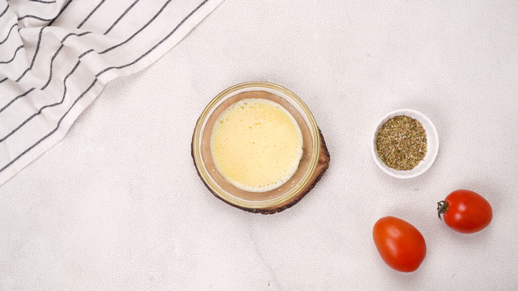 A small bowl of whisked egg wash on a wooden coaster, accompanied by a bowl of seasoning and fresh tomatoes, with a striped towel in the corner. Photo by Gluten-Free Palate.