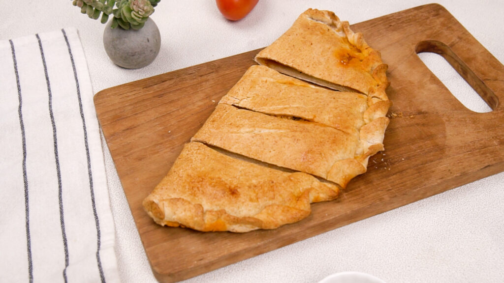 A freshly baked gluten-free calzone on a wooden cutting board, sliced into portions, with fresh tomatoes and a succulent plant nearby. Photo by Gluten-Free Palate.