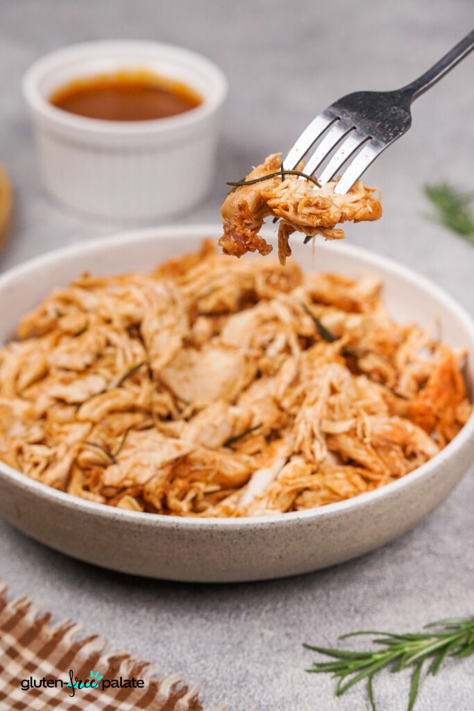 A fork lifting a saucy piece of shredded barbecue chicken over a bowl filled with more shredded chicken and rosemary sprigs.
