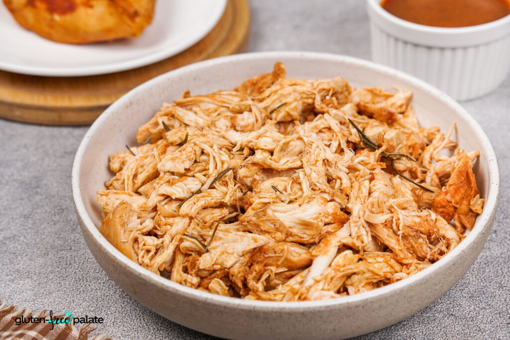 Instant Pot Shredded Barbecue Chicken A side view of a bowl filled with shredded barbecue chicken, served alongside extra BBQ sauce and chicken pieces on a plate.
