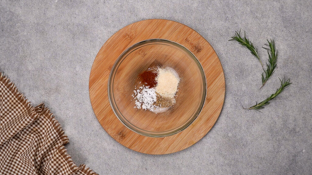 Step 1 A glass bowl with a mix of barbecue seasonings including garlic powder, paprika, and onion powder, resting on a wooden board with fresh rosemary nearby.