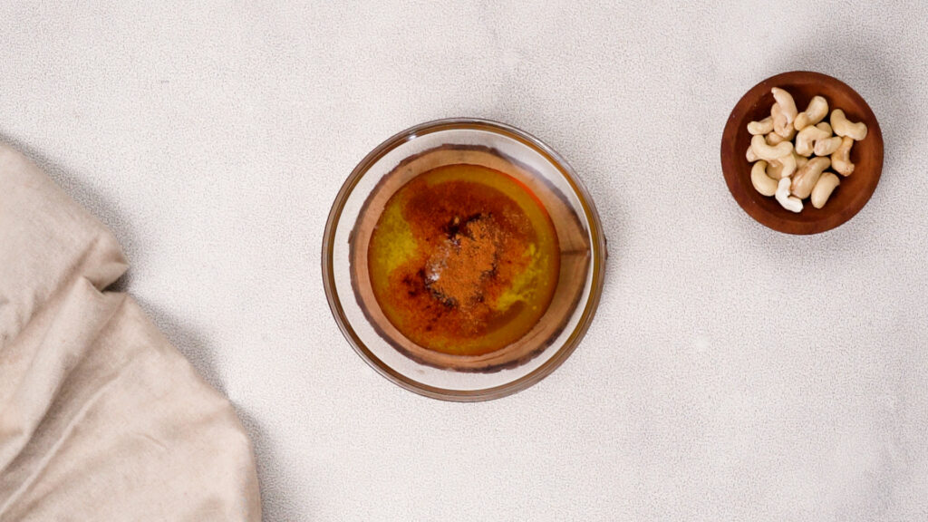 A glass bowl with melted butter, honey, and spices, ready to be mixed for honey-roasted cashews. A small wooden bowl of raw cashews sits nearby.