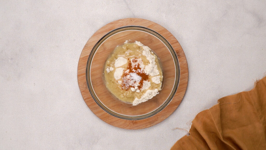 A glass bowl containing gluten-free masa harina, broth, spices, and baking powder on a wooden base.