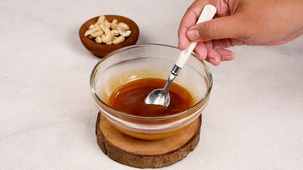 A hand stirring a glass bowl filled with a golden honey mixture, with a small wooden bowl of raw cashews in the background.