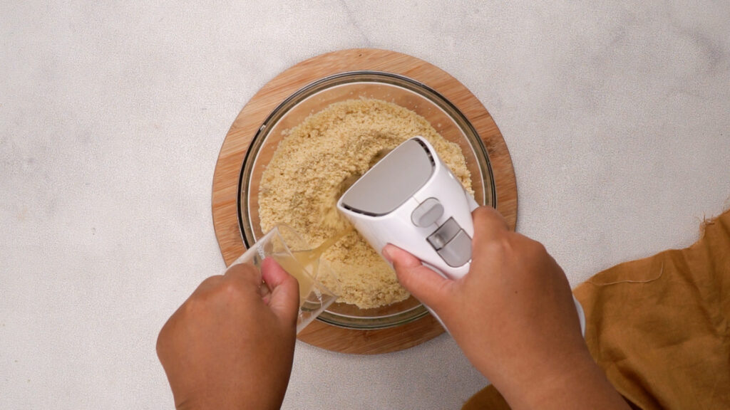 Hands using an electric mixer to combine masa dough and broth until smooth and fluffy.