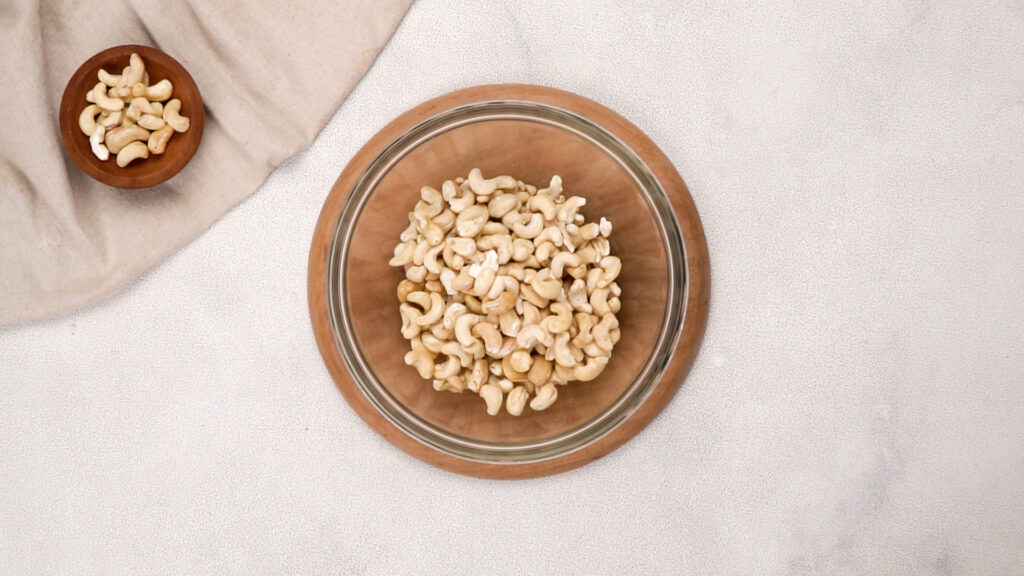 A glass bowl filled with raw cashews, placed on a wooden board, ready to be coated in a sweet honey glaze.