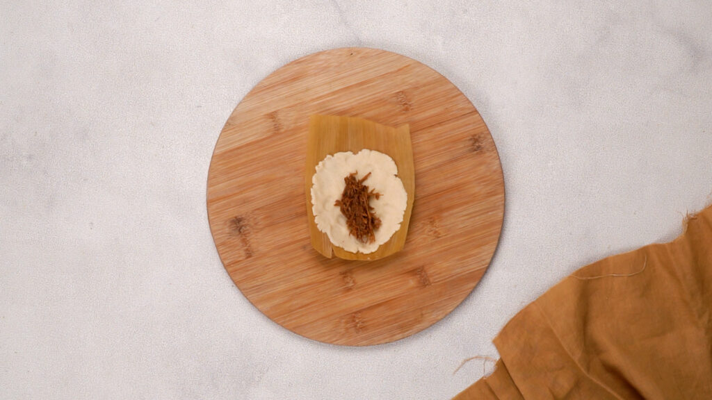 A corn husk with a layer of masa dough spread on it, topped with shredded meat filling.