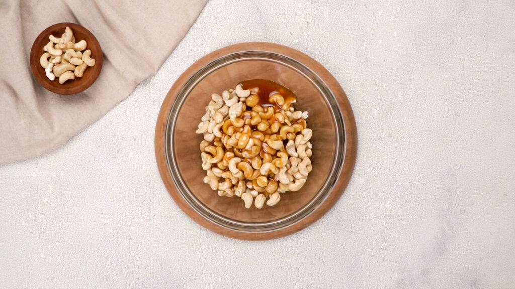 A glass bowl of raw cashews with freshly poured honey mixture on top, preparing to be stirred and coated evenly.