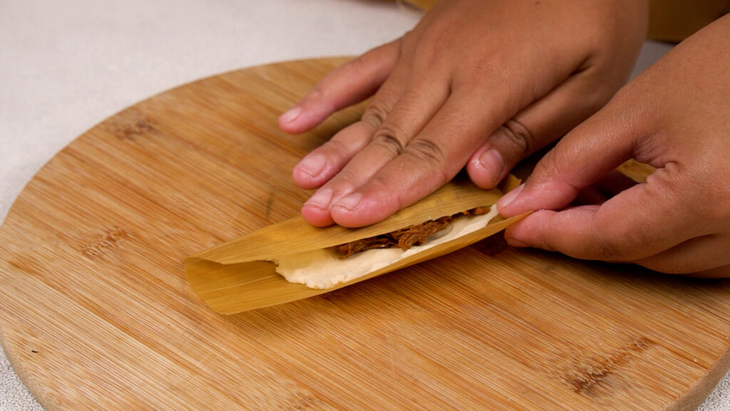 Hands carefully folding a corn husk over the masa and filling to form a tamale.