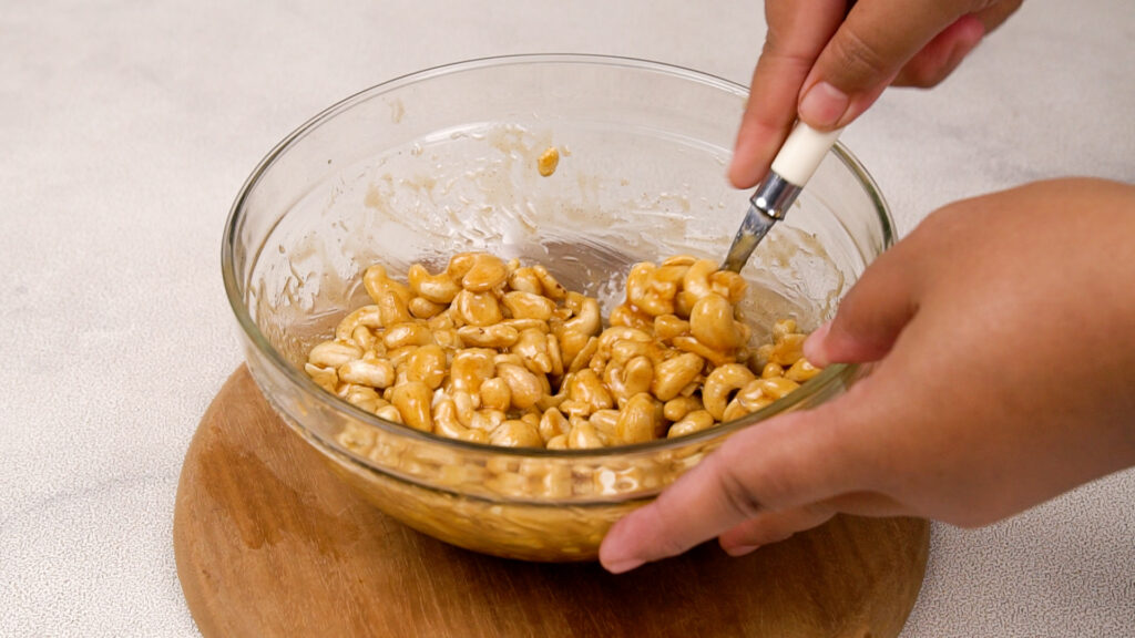 A hand mixing raw cashews with honey glaze in a glass bowl, ensuring each nut is evenly coated.