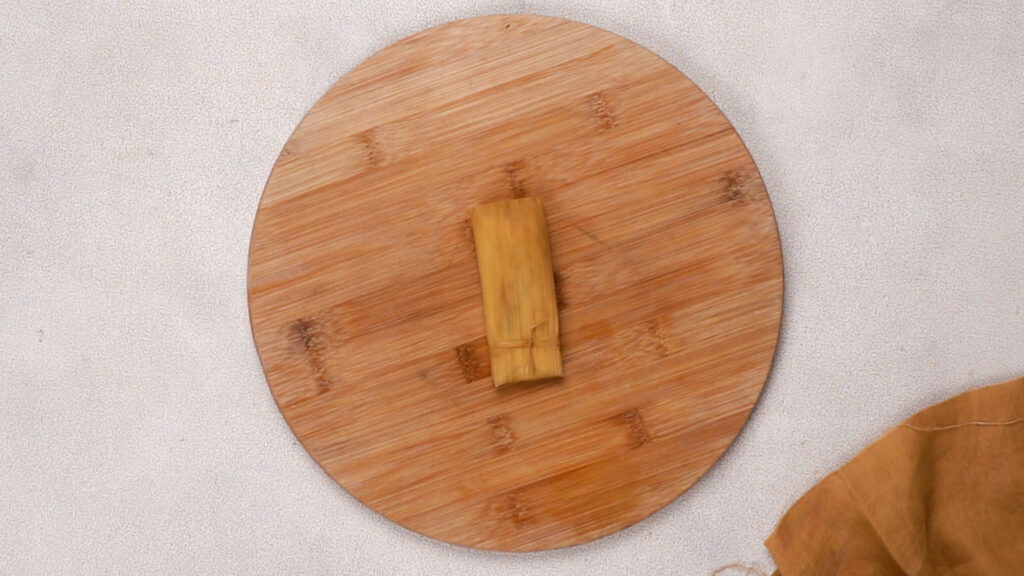 A fully wrapped and tied tamale resting on a wooden board, ready for cooking.