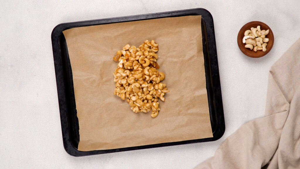 A parchment-lined baking tray with a mound of honey-coated cashews, ready to be spread and roasted.