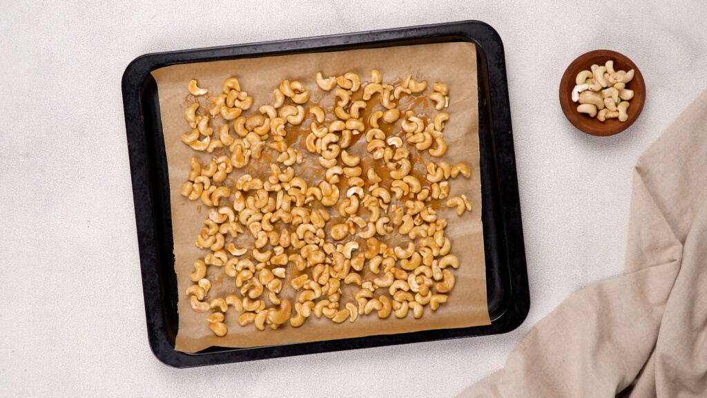 A baking tray with honey-coated cashews evenly spread on parchment paper, prepared for roasting.