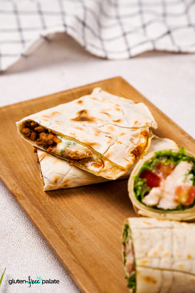 A close-up of gluten-free wrap with a golden-brown crust, filled with melted cheese, seasoned meat, and vegetables, cut into triangles and stacked on a wooden board. Beside them, a fresh chicken wrap with lettuce, tomato, and dressing is sliced in half.