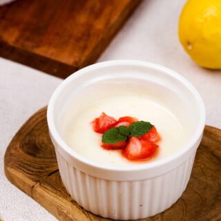 A close-up of a single white ramekin filled with lemon posset, garnished with diced strawberries and a sprig of fresh mint.