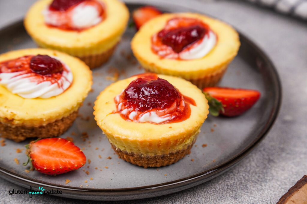 A plate of mini gluten-free cheesecakes with golden crusts, topped with strawberry sauce and cream, garnished with fresh strawberry slices..