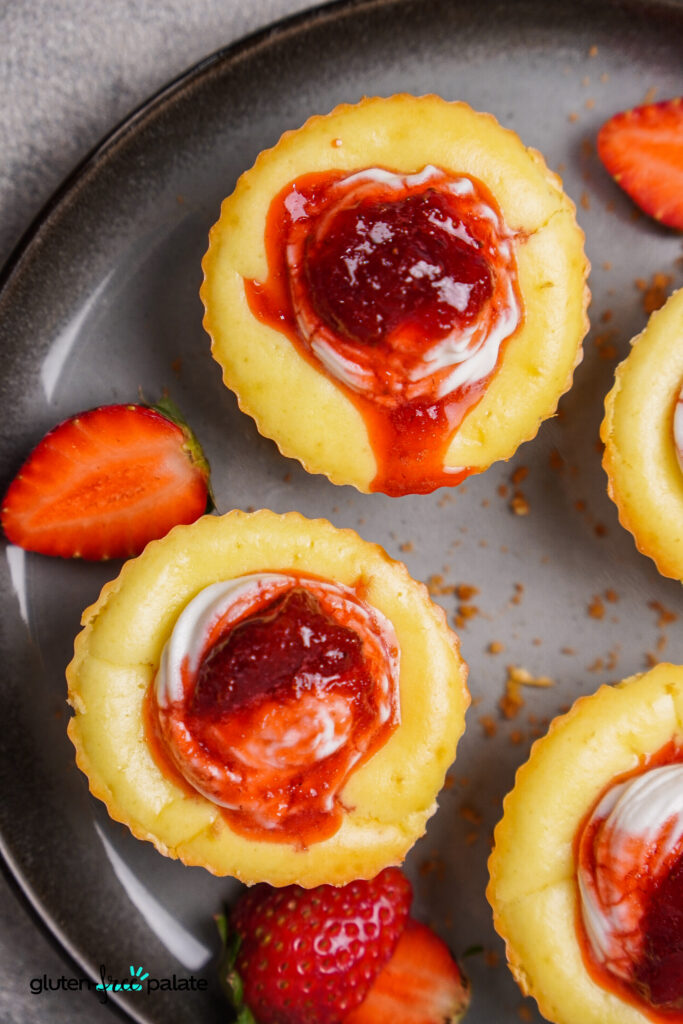 Close-up of mini gluten-free cheesecake bites topped with strawberry sauce and a dollop of sour cream, served on a dark plate with fresh strawberry slices.