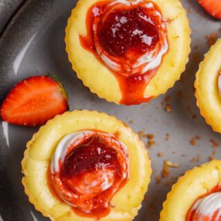 Close-up of mini gluten-free cheesecake bites topped with strawberry sauce and a dollop of sour cream, served on a dark plate with fresh strawberry slices.