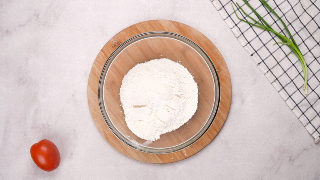 A glass bowl filled with gluten-free flour sits on a round wooden board, with a fresh tomato and green onions nearby.