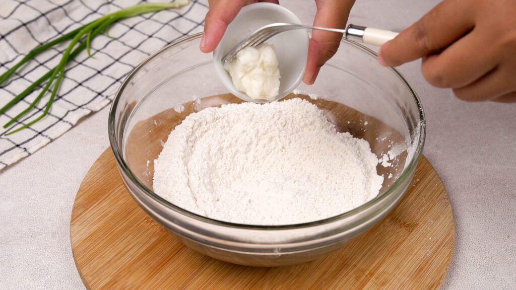 A hand adding in solid fat into a glass bowl filled with gluten-free flour, preparing to mix the ingredients for a soft and pliable dough.