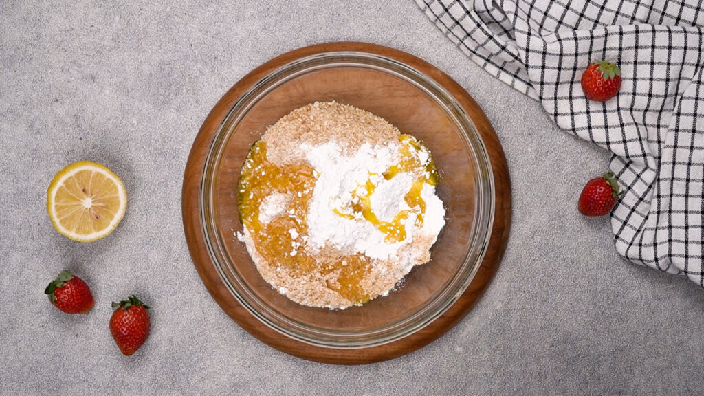 A glass bowl filled with crushed graham crackers, powdered sugar, and melted butter, ready to be mixed for the cheesecake crust.