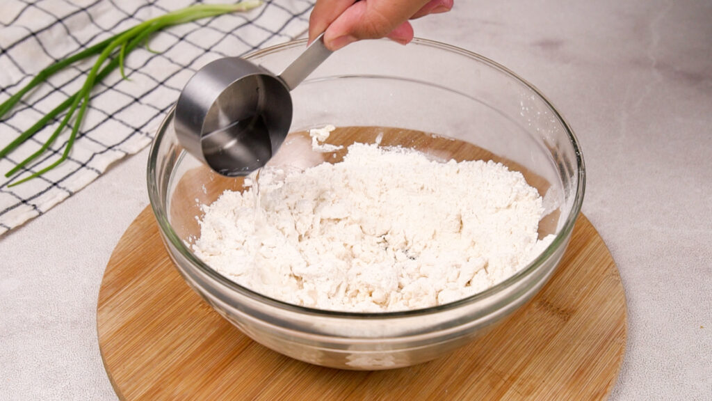 A measuring cup pours water into a bowl of gluten-free flour mixture, beginning the process of forming the dough.