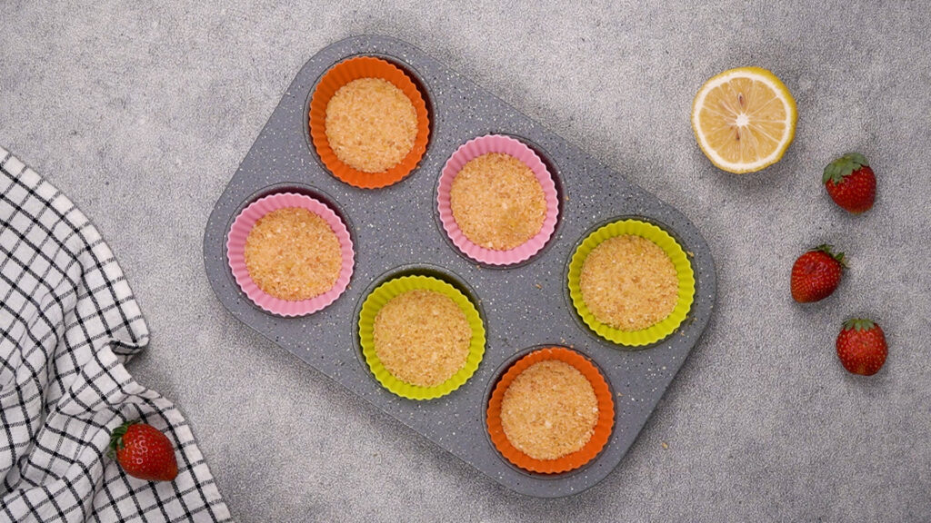 A muffin tin with pressed gluten-free graham cracker crusts in silicone baking cups.