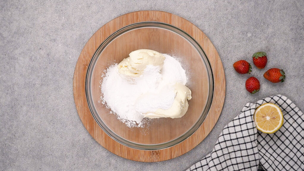 A glass mixing bowl containing softened cream cheese and powdered sugar, ready to be blended into a creamy cheesecake filling.