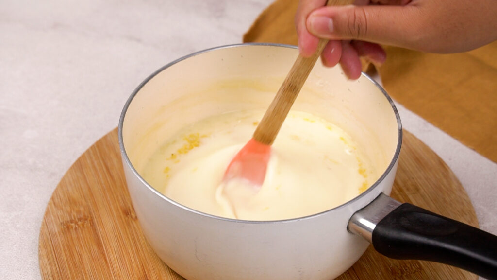 A hand stirs the lemon zest into the cream mixture with a spatula.