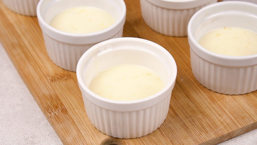 The creamy lemon mixture is poured into individual white ramekins placed on a wooden board.