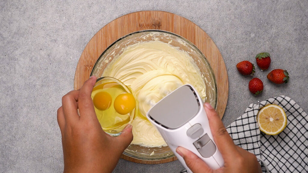 A hand mixer blending cheesecake batter while eggs are being added.