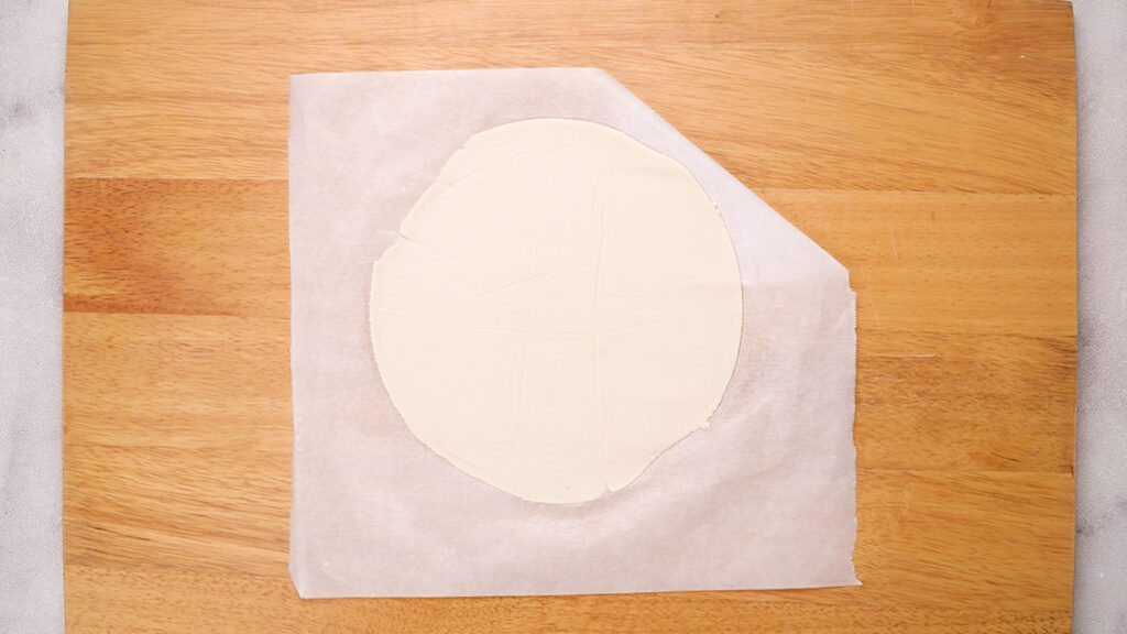 A freshly rolled-out gluten-free tortilla sits on parchment paper atop a wooden cutting board, shaped into a thin, round wrap.