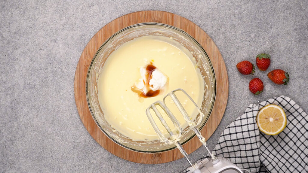 A mixing bowl with smooth cheesecake batter, sour cream, and vanilla extract being added before final blending.