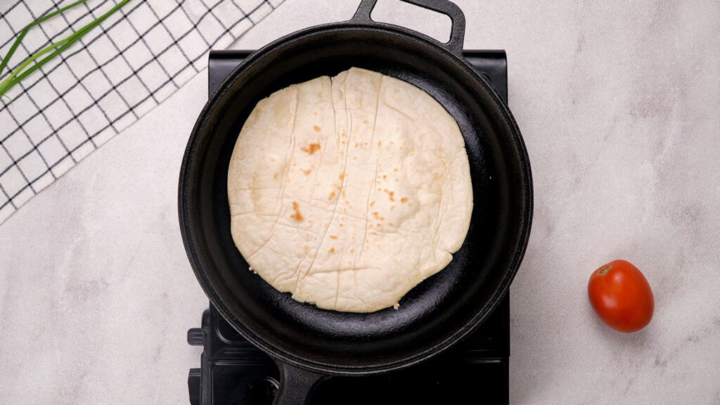 A gluten-free wrap cooking in a black cast iron pan, developing light golden brown spots on its surface.
