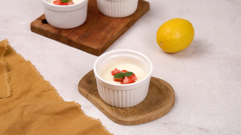 A garnished lemon posset topped with diced strawberries and a mint sprig in a ramekin on a small wooden board.