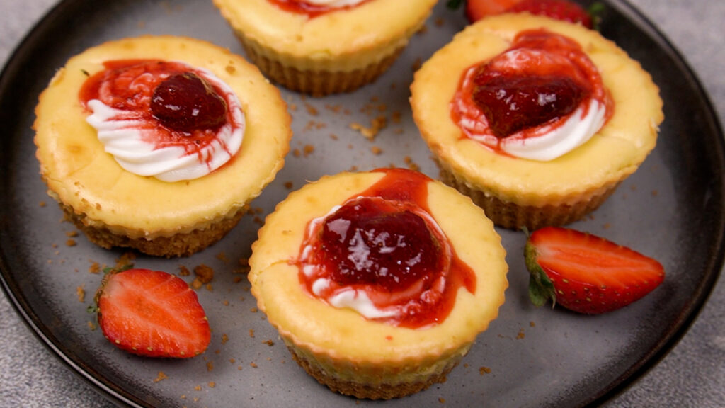 Mini gluten-free cheesecakes on a dark plate, topped with swirls of cream and strawberry sauce, garnished with fresh strawberries.