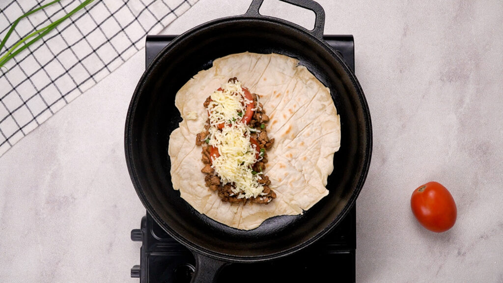A gluten-freewrap in a cast iron skillet topped with seasoned meat, sliced tomatoes, and shredded cheese, cooking to melt the cheese for a flavorful filling.