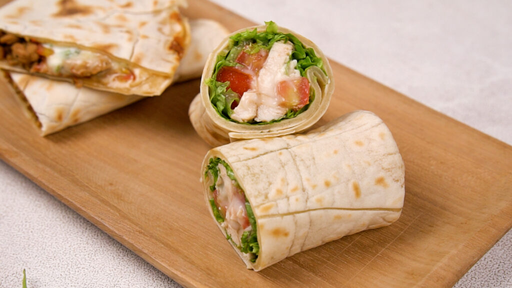 A finished gluten-free wrap filled with fresh greens, tomatoes, chicken, and dressing is cut in half and placed on a wooden board for serving.