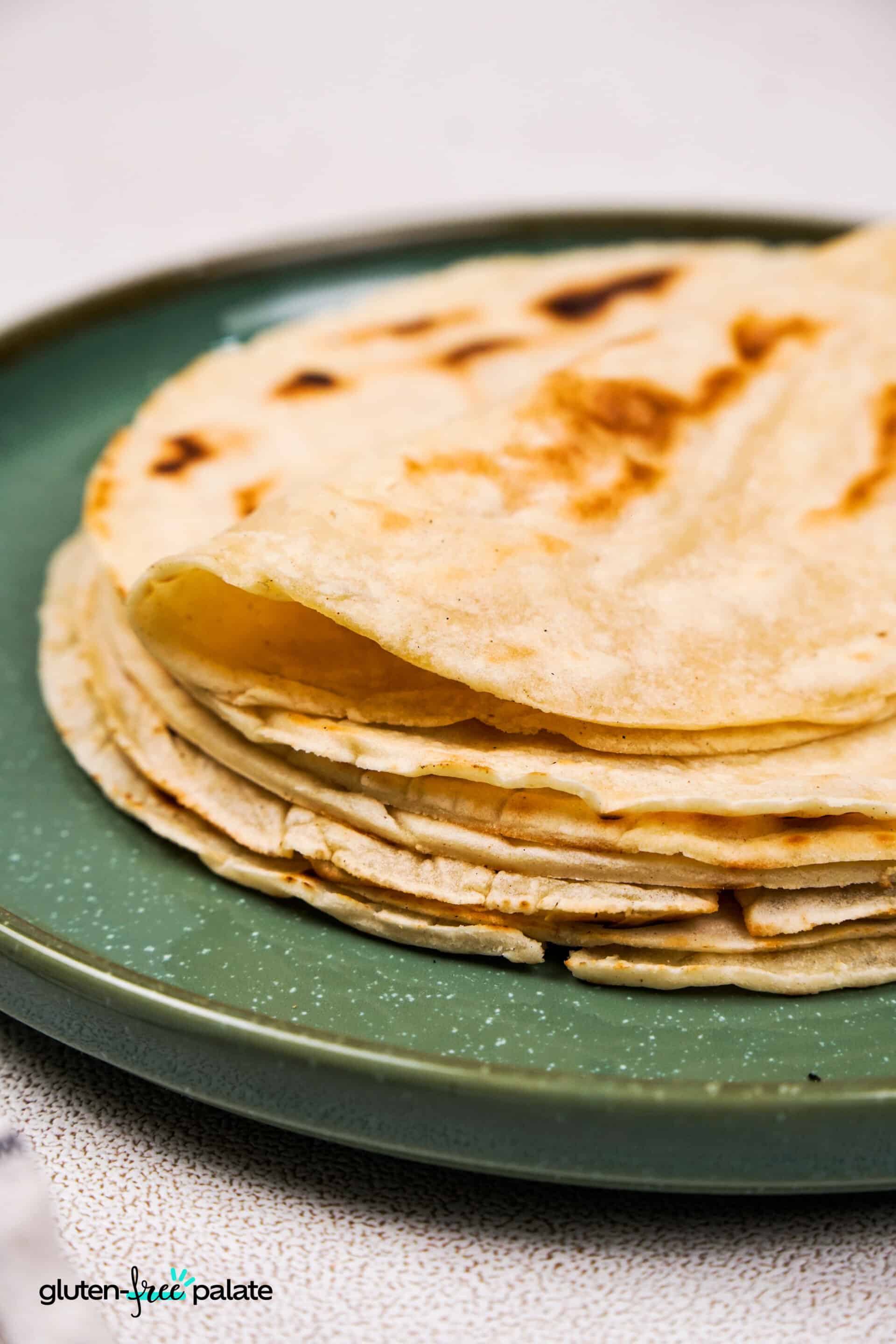 Close-up of a stack of gluten-free tacos on a green ceramic plate, showcasing their soft, foldable texture and golden brown spots.