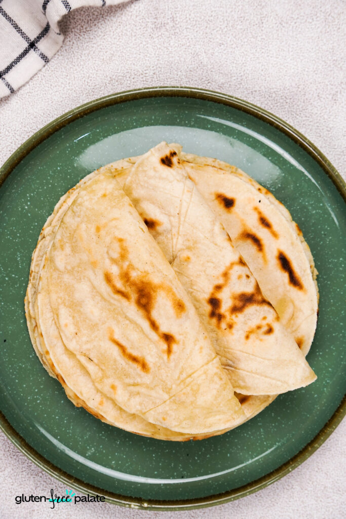 An overhead shot of folded gluten-free tacos stacked neatly on a green plate, with a checkered napkin in the corner.