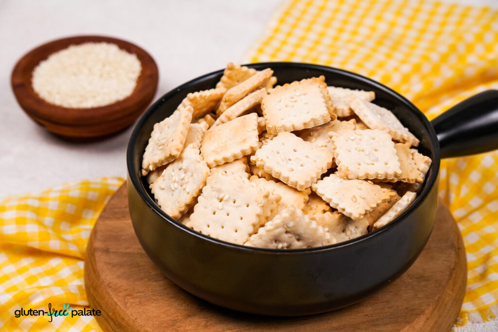 A black bowl filled with square, golden-brown gluten-free crackers topped with sesame seeds, placed on a wooden board with a yellow checkered cloth and a small wooden bowl of sesame seeds in the background.