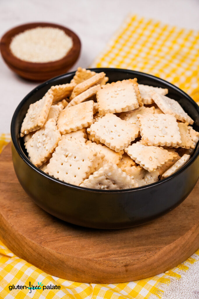 A top-down view of a bowl filled with crispy gluten-free crackers, with sesame seeds sprinkled on top and a yellow gingham cloth underneath.
