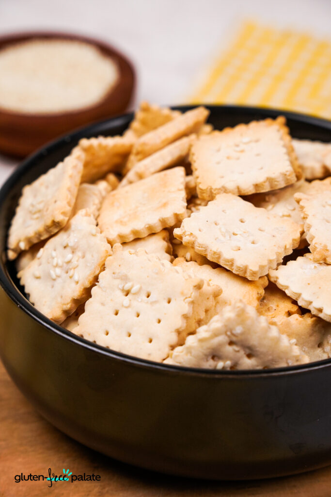 A close-up view of a bowl full of golden, square gluten-free crackers with scalloped edges and sesame seeds on top, placed beside a wooden bowl of sesame seeds.