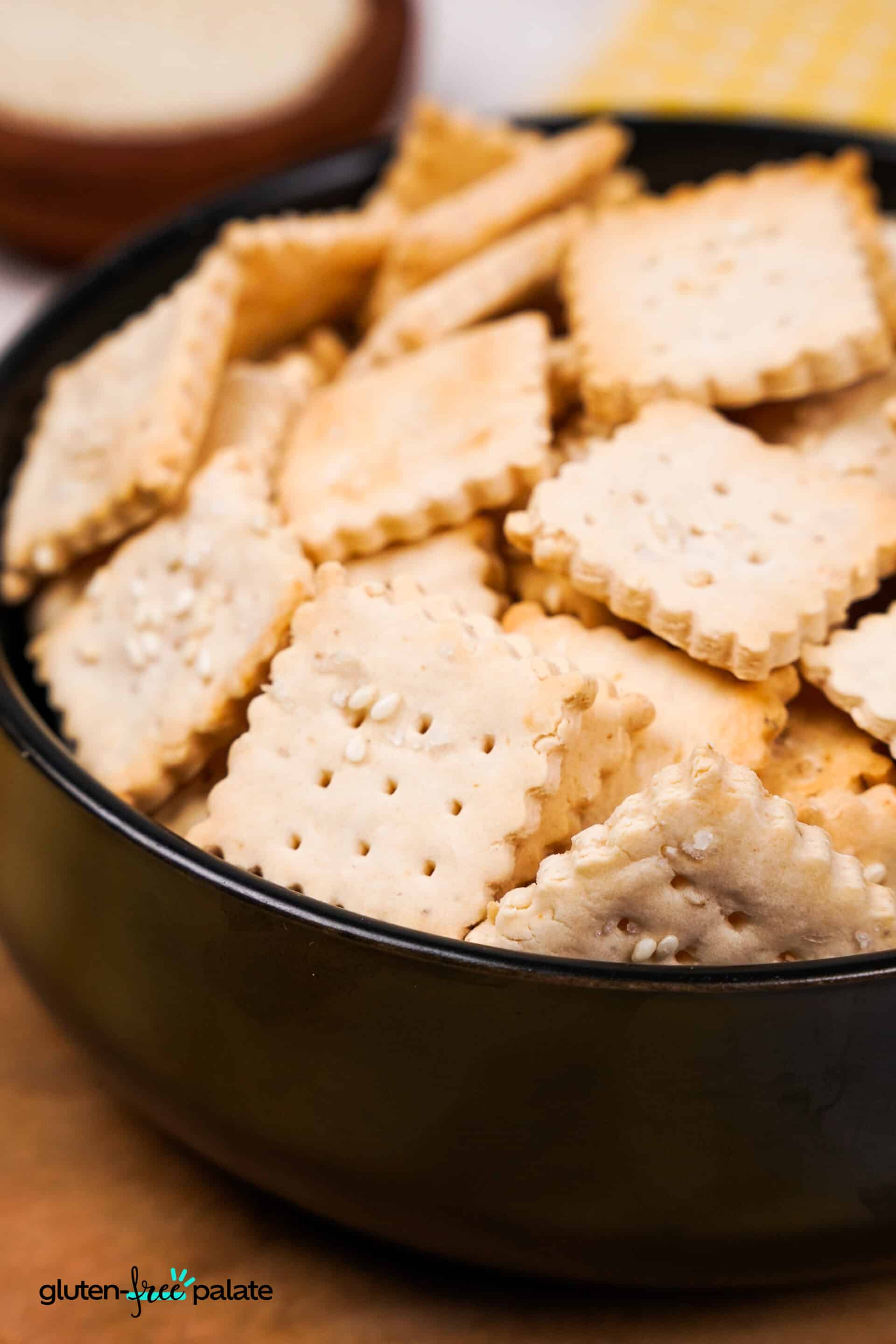 A side-angle view of a black bowl filled with square gluten-free crackers, showing their crisp texture and sesame seed topping, with part of a yellow gingham cloth visible in the background.
