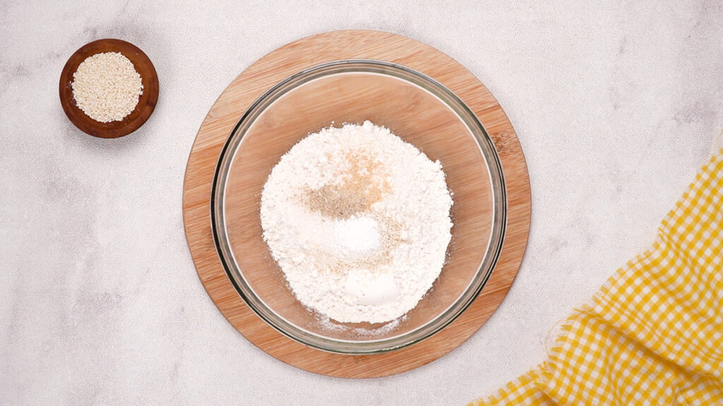 A glass mixing bowl filled with gluten-free flour, sesame seeds, garlic powder, baking powder, and other dry ingredients, ready to be stirred.