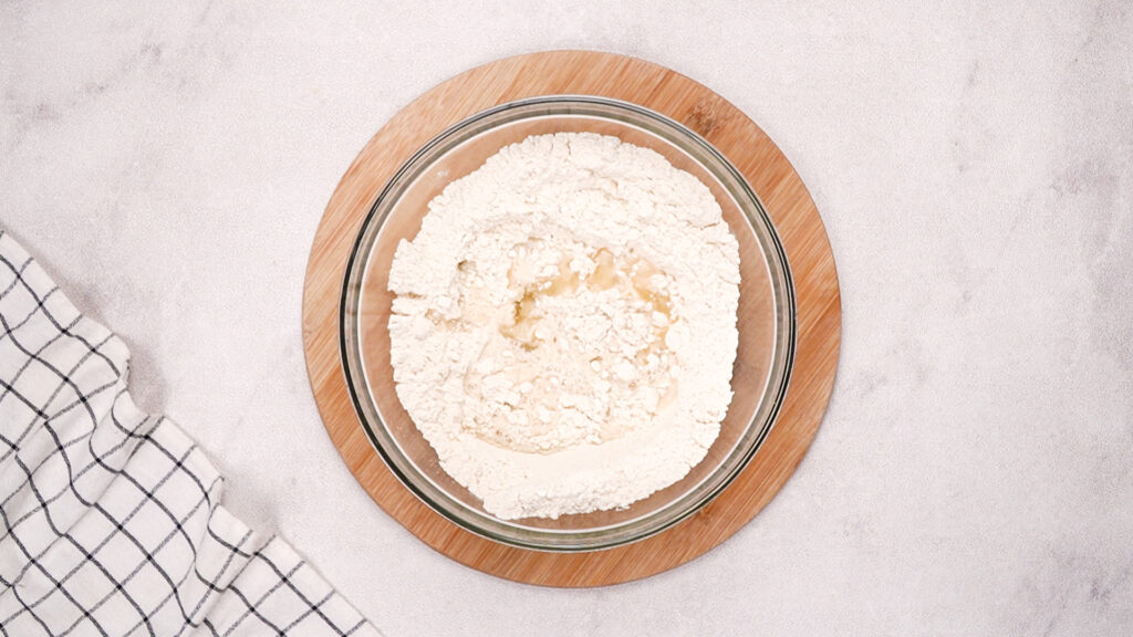 Dry ingredients in the glass bowl with water and oil just added, ready to be mixed into dough.