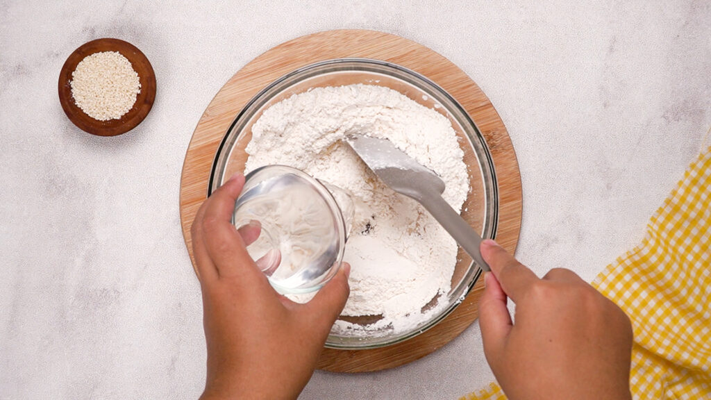 A hand pouring water from a glass cup into the flour mixture while another hand holds a spatula, starting to form the gluten-free cracker dough.