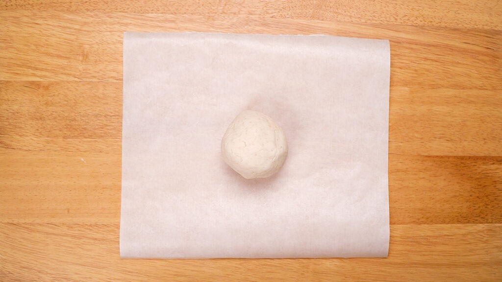 A ball of gluten-free cracker dough placed between two sheets of parchment paper on a wooden surface, prepared for rolling out.