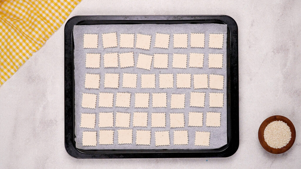 A parchment-lined baking sheet filled with rows of square gluten-free crackers, ready to go into the oven.