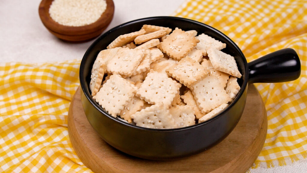 A black bowl filled with golden-brown, square gluten-free crackers topped with sesame seeds, sitting on a wooden board with a yellow gingham cloth.